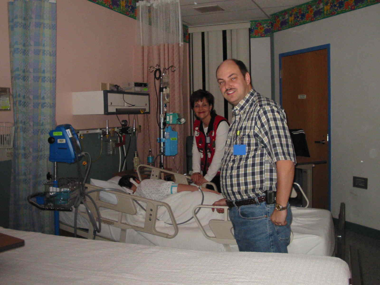 Isaac resting in a hospital bed while Doug and Marsha stand by the side of the bed